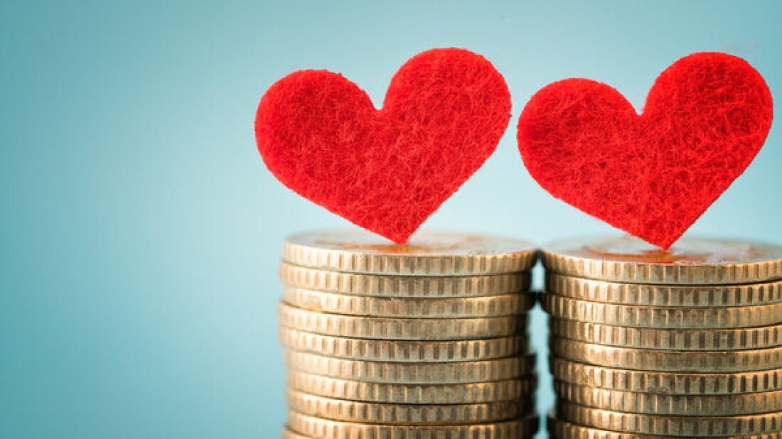 Two felt red hearts are balanced on two separate stacks of gold coins on a light blue background, representing people in a romantic relationship that brings finances together.